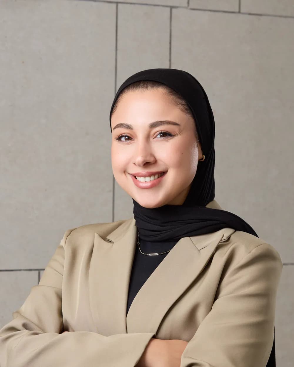 Farah Al-Holaysi in a beige blazer and black headscarf, arms crossed, against a stone-tiled wall