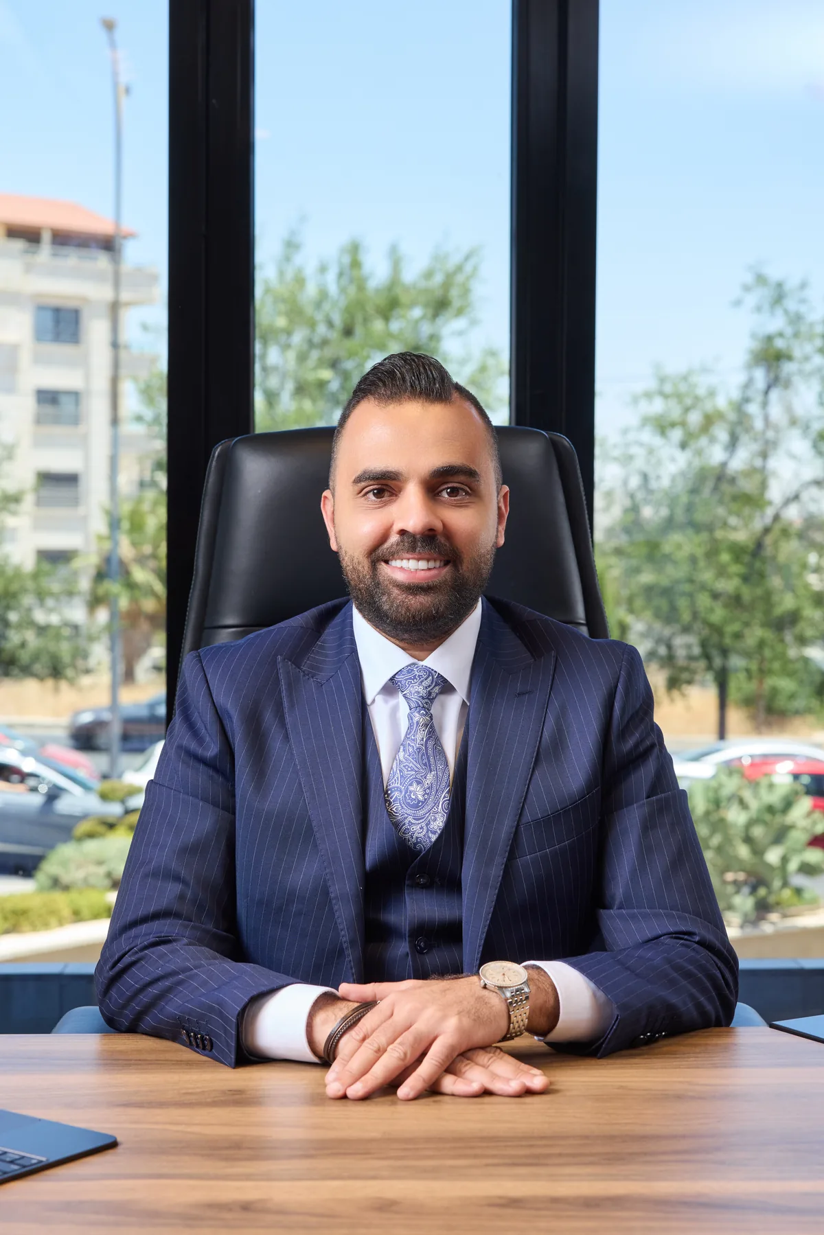 Rami Musallam in a navy pinstripe three-piece suit, seated at a desk in front of floor-to-ceiling office windows